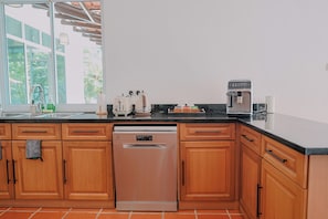 A kitchen with wooden cabinets, a dishwasher, a coffee maker, and a window with a view of greenery. - Villa 7 - Luxury Private Pool Villa (Langkawi)