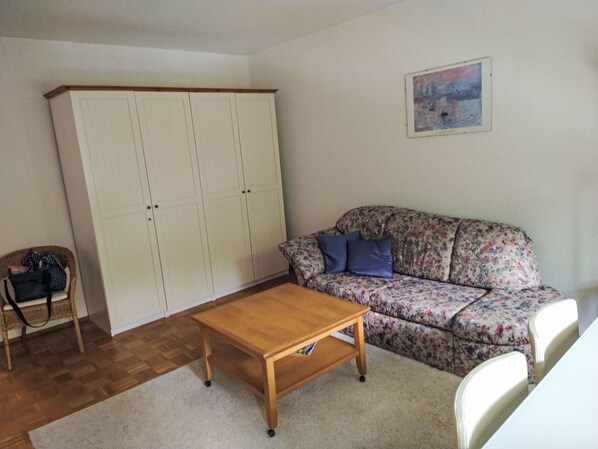 A room with a wooden table, a floral sofa, a white wardrobe, and a wicker chair.