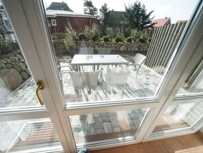Eine Glastür führt zu einer Terrasse im Freien mit Tisch und Stühlen, einer Steinsäule und Blick auf Häuser und Bäume.