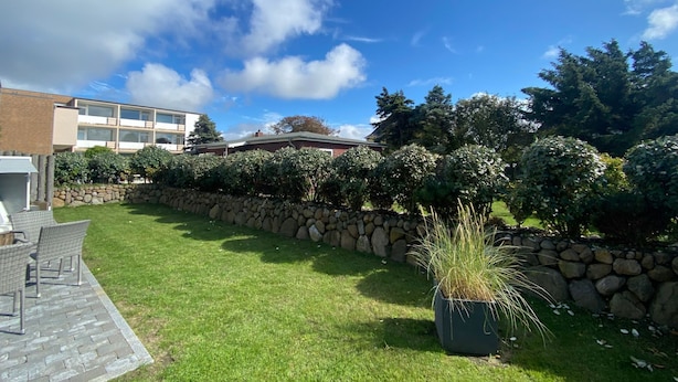 Un patio con muro de piedra, zona verde y área de descanso con sillas.