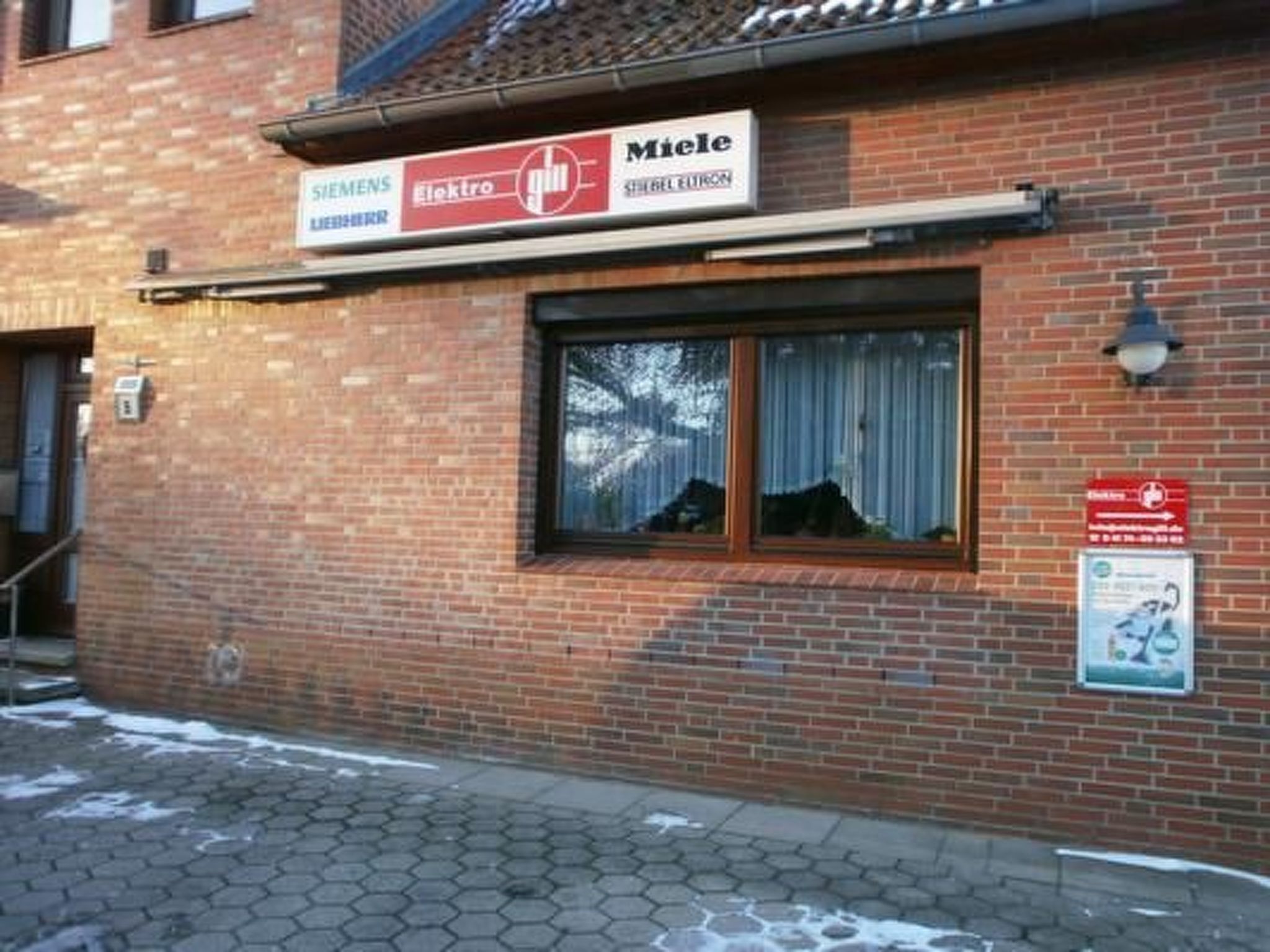 Ein Backsteinhaus mit Schildern von Siemens und Miele, einem Fenster mit Vorhängen und einem roten Notausgangsschild.