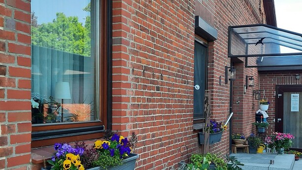 Un edificio de ladrillo con macetas de flores en la planta baja, una ventana con vista a los árboles, y una puerta con un letrero encima.