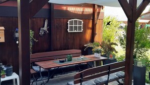 Una terraza de madera con una mesa y bancos, rodeada de plantas en macetas y una valla de madera.