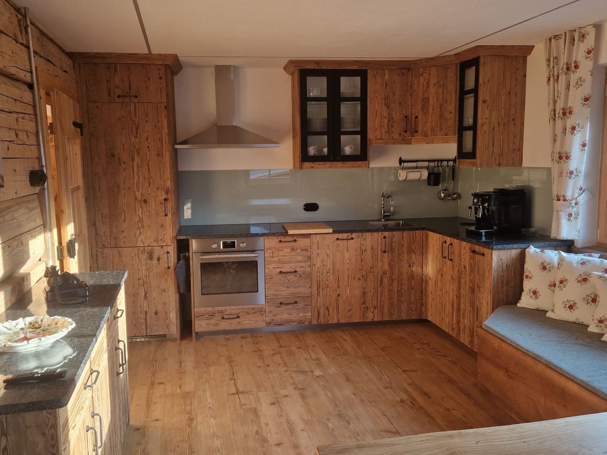 Una cucina compatta con mobili in legno, forno a incasso, macchina da caffè e una finestra con tende floreali.