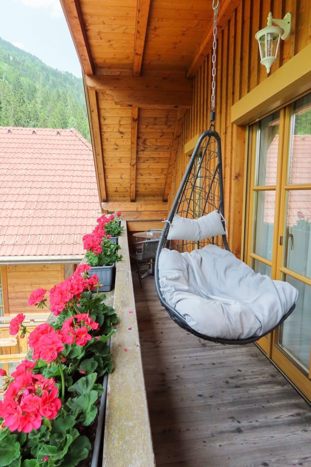 A wooden balcony with a hanging chair, potted geraniums, and a view of a red-tiled roof building.