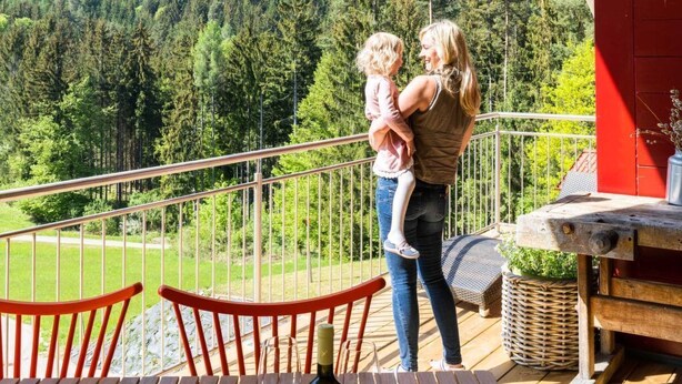 Ein Balkon mit Blick auf eine bewaldete Berglandschaft, einem Holztisch mit Weingläsern und einer Flasche sowie einem roten Stuhl.