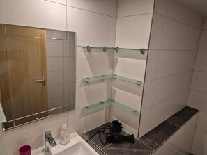 Un bany amb una porta de fusta, un mirall, un lavabo i tres prestatges de vidre.