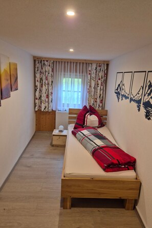 Un dormitori petit i acollidor amb un llit de fusta, una tauleta de nit, una finestra amb cortines florals i decoració mural amb motius de muntanya.