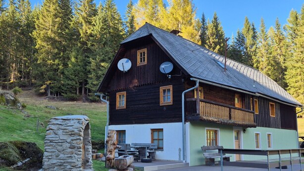 Ein zweistöckiges Holzhaus mit Satellitenschüsseln, einer Steinmauer und einer kleinen Sitzgelegenheit im Freien.