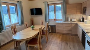 Eine moderne Küche mit Holzschränken, einem Esstisch und Stühlen. Es gibt Fenster mit Vorhängen, einen an der Wand montierten Fernseher und einen eingebauten Backofen.