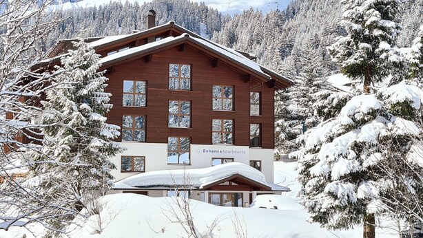 A wooden chalet with multiple windows and a sign reading "Bohmemia Apartments" amidst a snowy mountain landscape. - Bohemia 3 in Gargellen by Interhome (Gargellen)