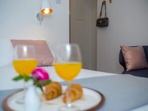 Una cama bien hecha con almohadas, una lámpara de noche, una mesita con cruasanes y un vaso de zumo de naranja, y un bolso colgado en la pared.
