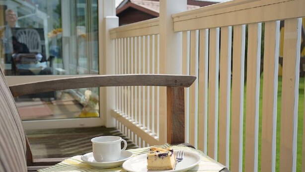 Een balkon met een houten stoel, een witte kop, een bord met een stuk taart en een geruit placemat.