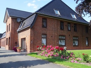 A large brick building with a steeply pitched roof, multiple windows, and a well-maintained garden with colorful flowers. - Modern eingerichtete Suite mit Terrasse by Interhome (Elskop)