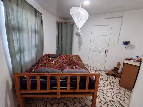 Une chambre avec un lit en bois, une couvre-lit à motifs, une moustiquaire, une porte blanche et une petite armoire en bois.