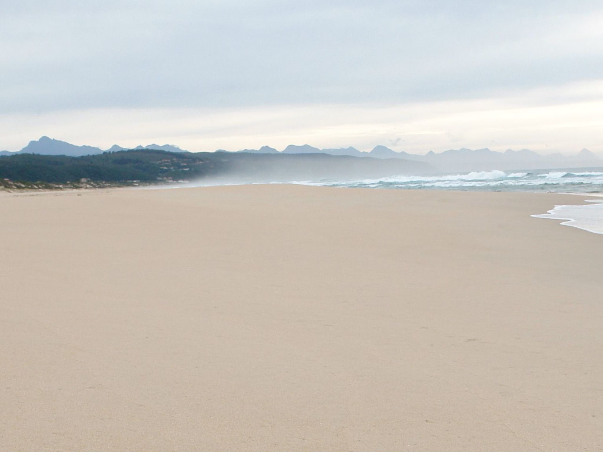 Una playa extensa y arenosa con olas suaves y una cadena montañosa al fondo.