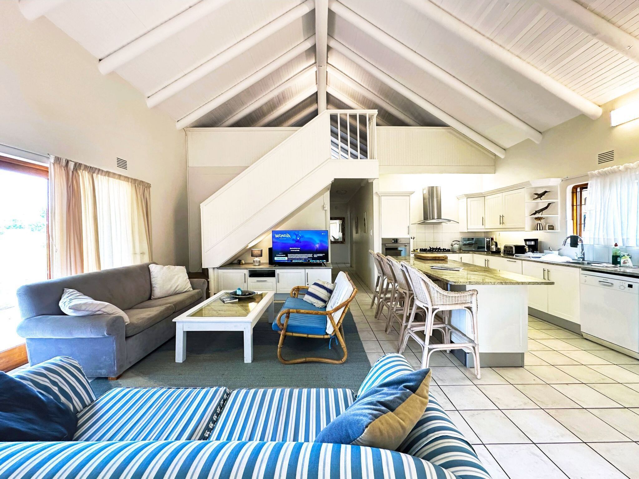 A spacious living area with a sofa, a coffee table, and a TV. There is a kitchen with a bar and a staircase leading to an upper level.