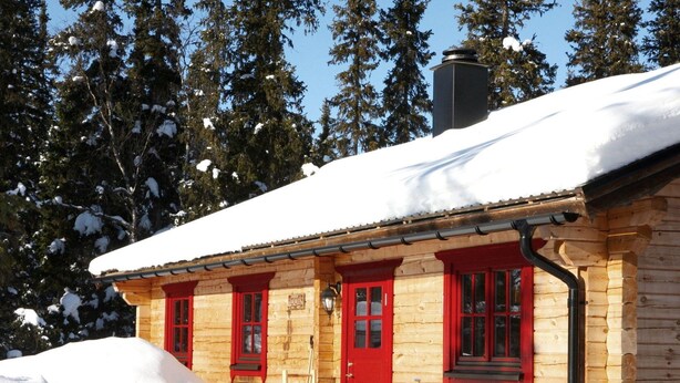 A wooden cabin with red doors and windows, surrounded by snow-covered trees.