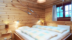 A wooden cabin bedroom with a bed, two bedside lamps, a hanging light fixture, and a window with curtains.
