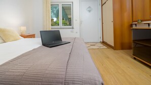 Un dormitorio con cama, una laptop sobre la cama, una ventana con cortinas, un armario y una mesita de noche con lámpara.