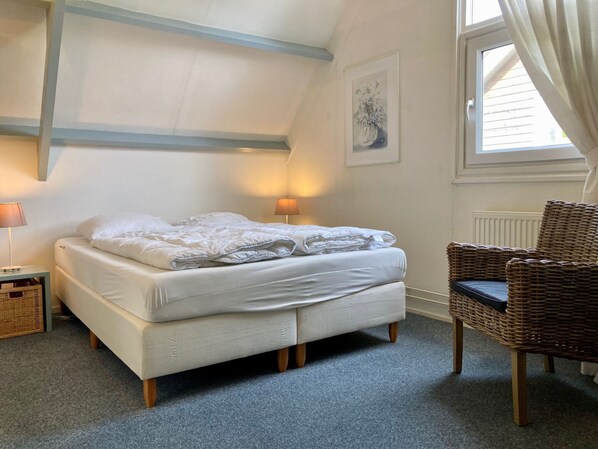 A bedroom with a bed, two bedside lamps, a wicker chair, a framed artwork, and a window with curtains.