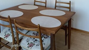 Ein Essbereich mit einem Holztisch und vier Stühlen, jeweils mit einem floralen Kissen. Über dem Tisch hängen drei gerahmte Bilder.