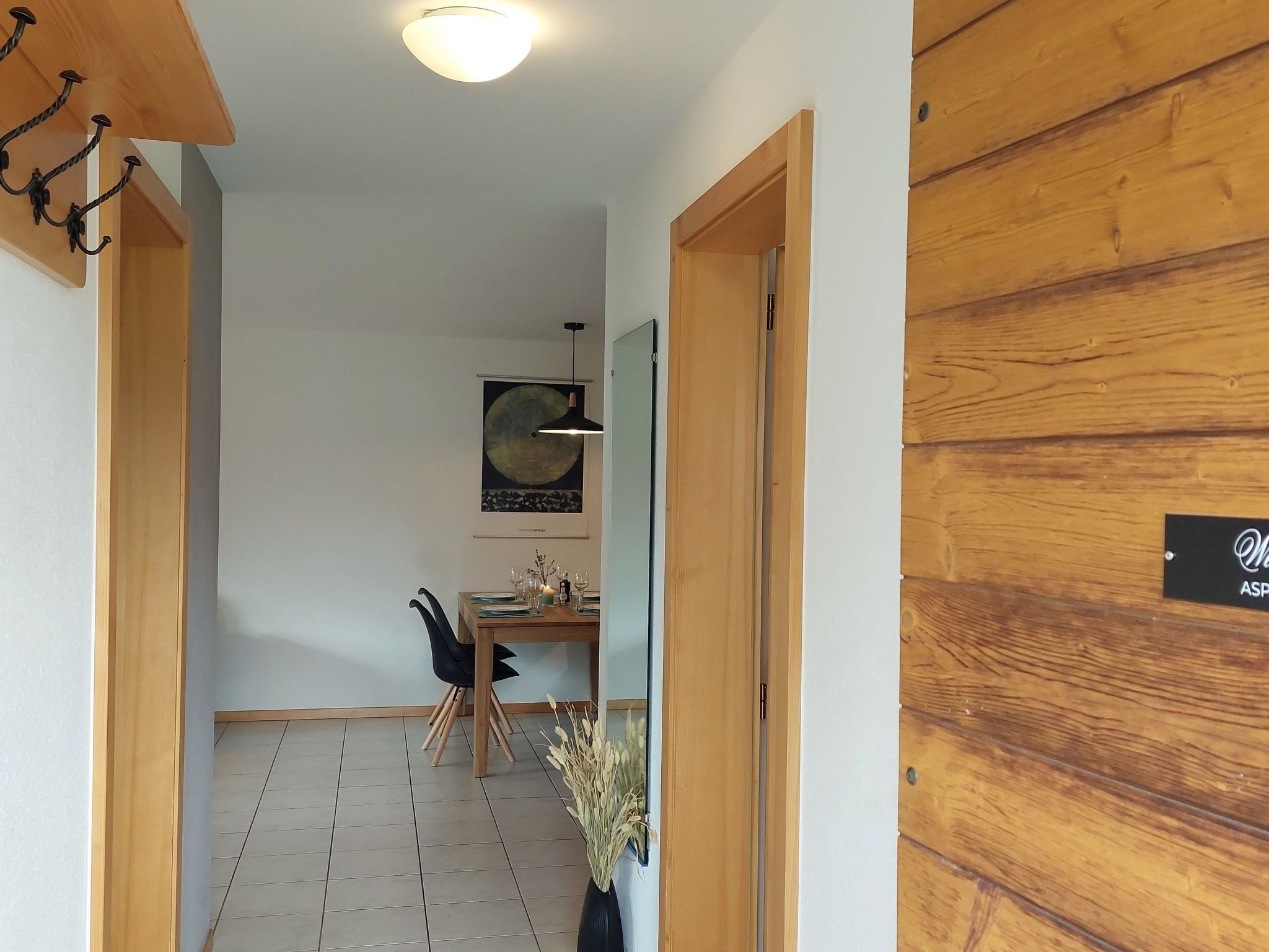 Un pasillo con una puerta de madera, un comedor con mesa y sillas, y un aplique en la pared.