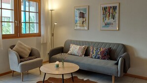 Ett vardagsrum med en grå soffa, en liten fåtölj, ett runt soffbord med ett ljus och två inramade tavlor på väggen.
