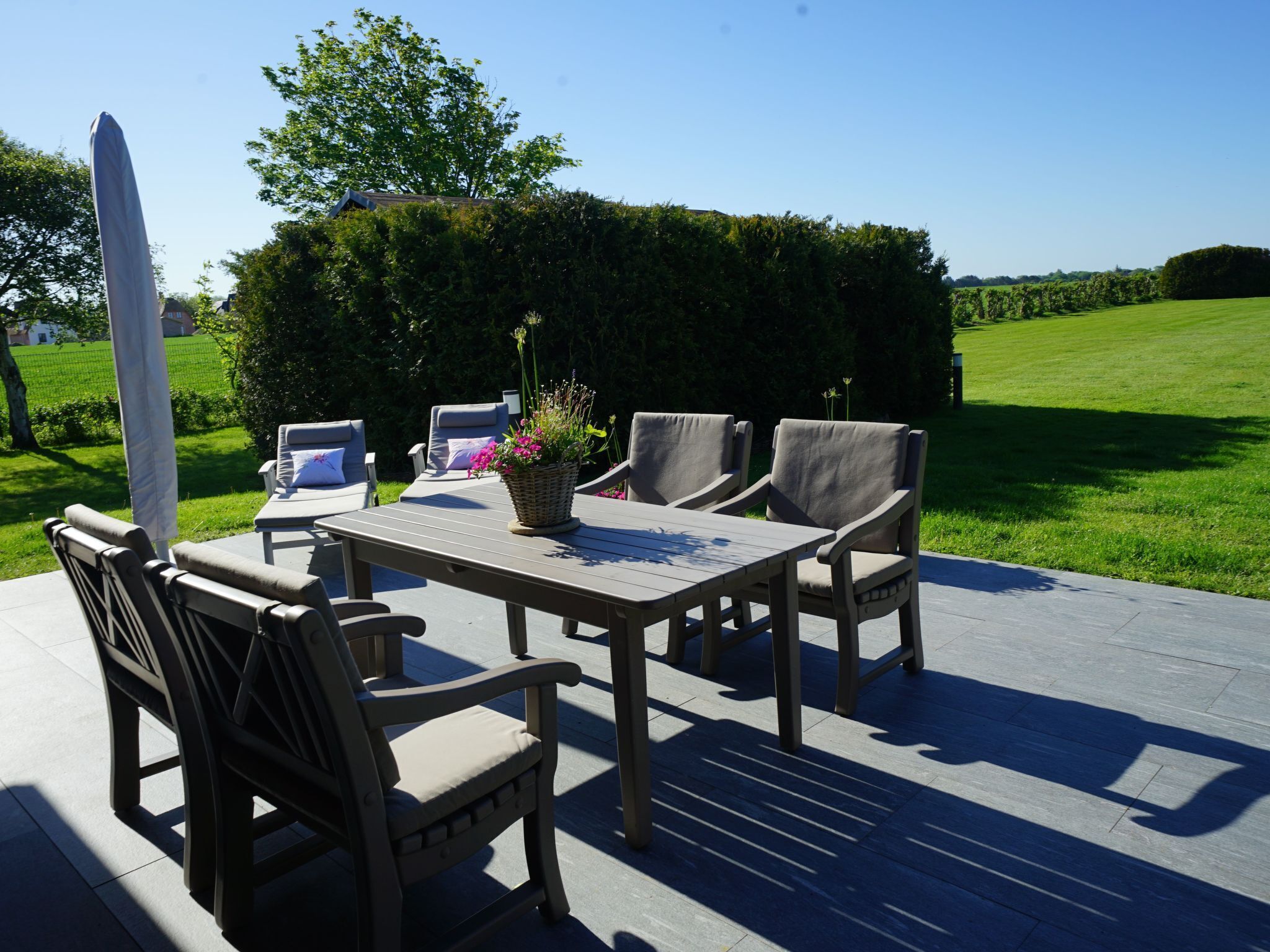 An outdoor patio with a table and chairs, surrounded by a hedge and a lawn.