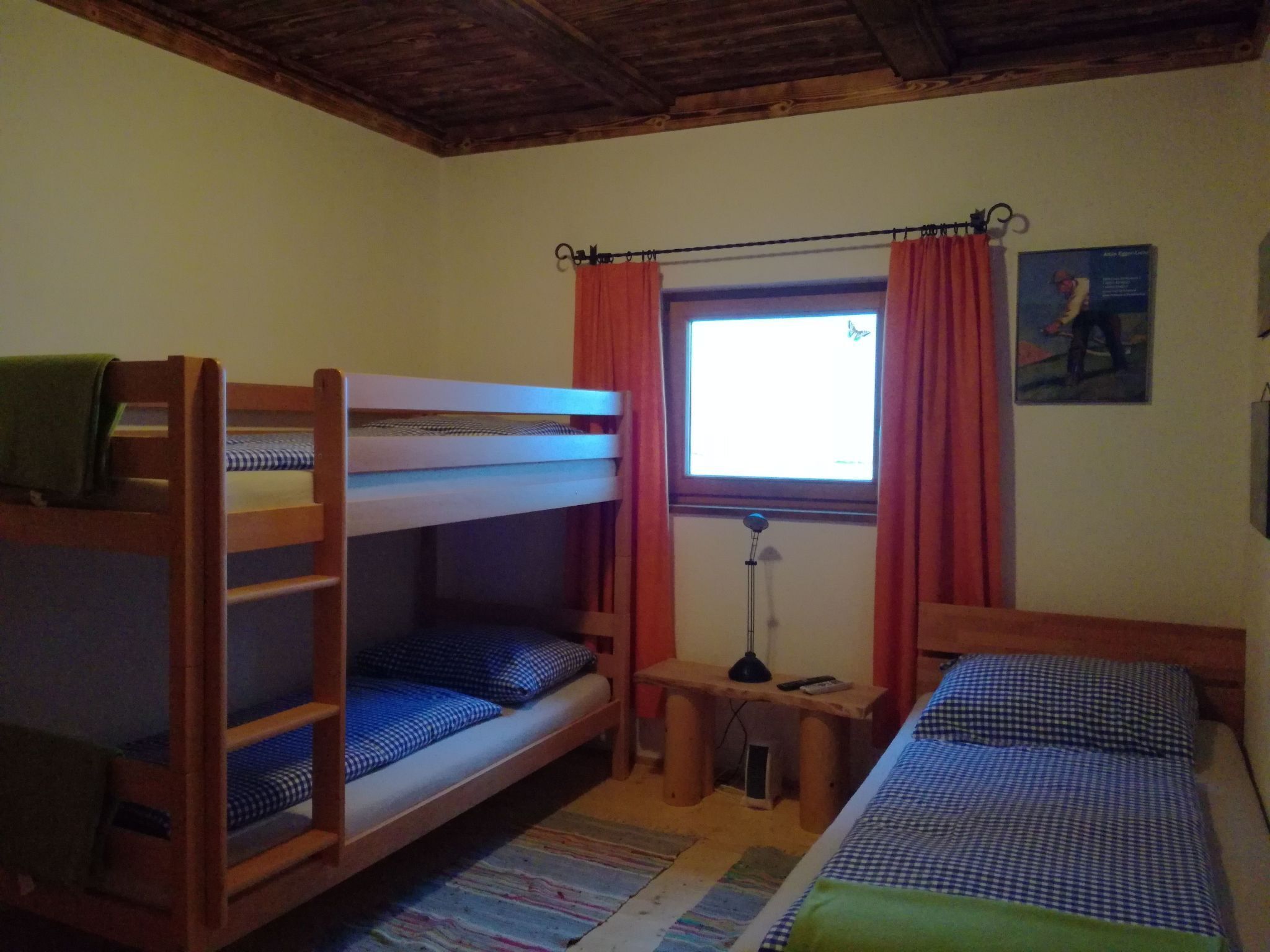A room with bunk beds, a small table with a lamp, a window with curtains, and a painting on the wall.