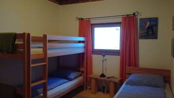 A room with bunk beds, a small table with a lamp, a window with curtains, and a painting on the wall.