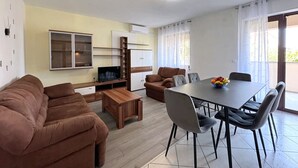 Een moderne woonkamer met een bruine bank, een houten salontafel en een televisie. Er is een eethoek met een tafel en stoelen, en een keuken met kasten en een televisie.