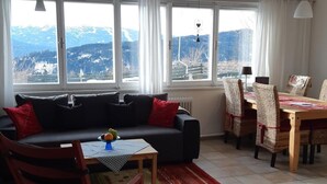 Een woonkamer met een bank, een eettafel met stoelen en uitzicht op bergen door grote ramen.