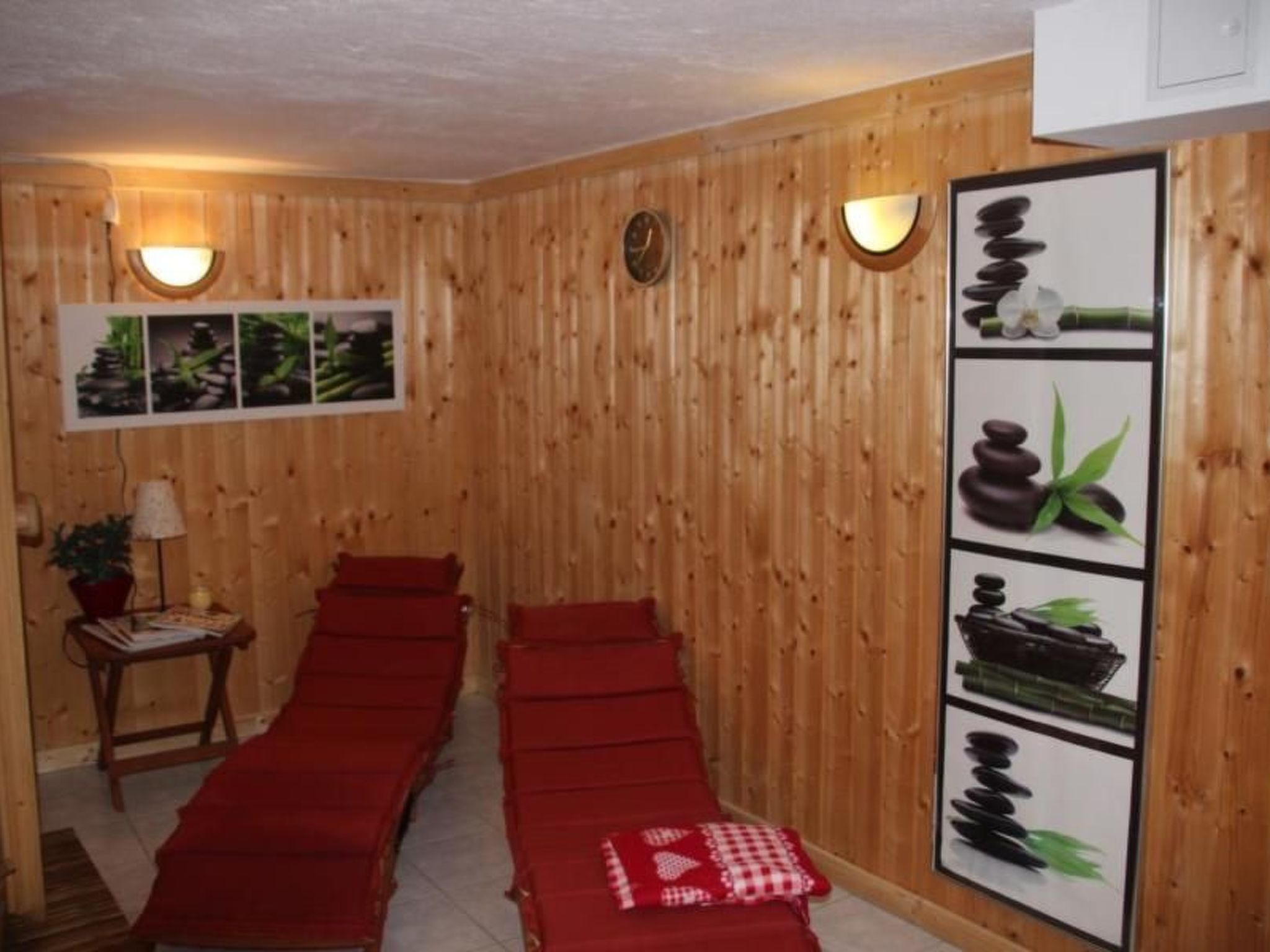 Une pièce aux murs lambrés, avec deux tables de massage rouges, une horloge murale et un tableau représentant des pierres et des plantes.