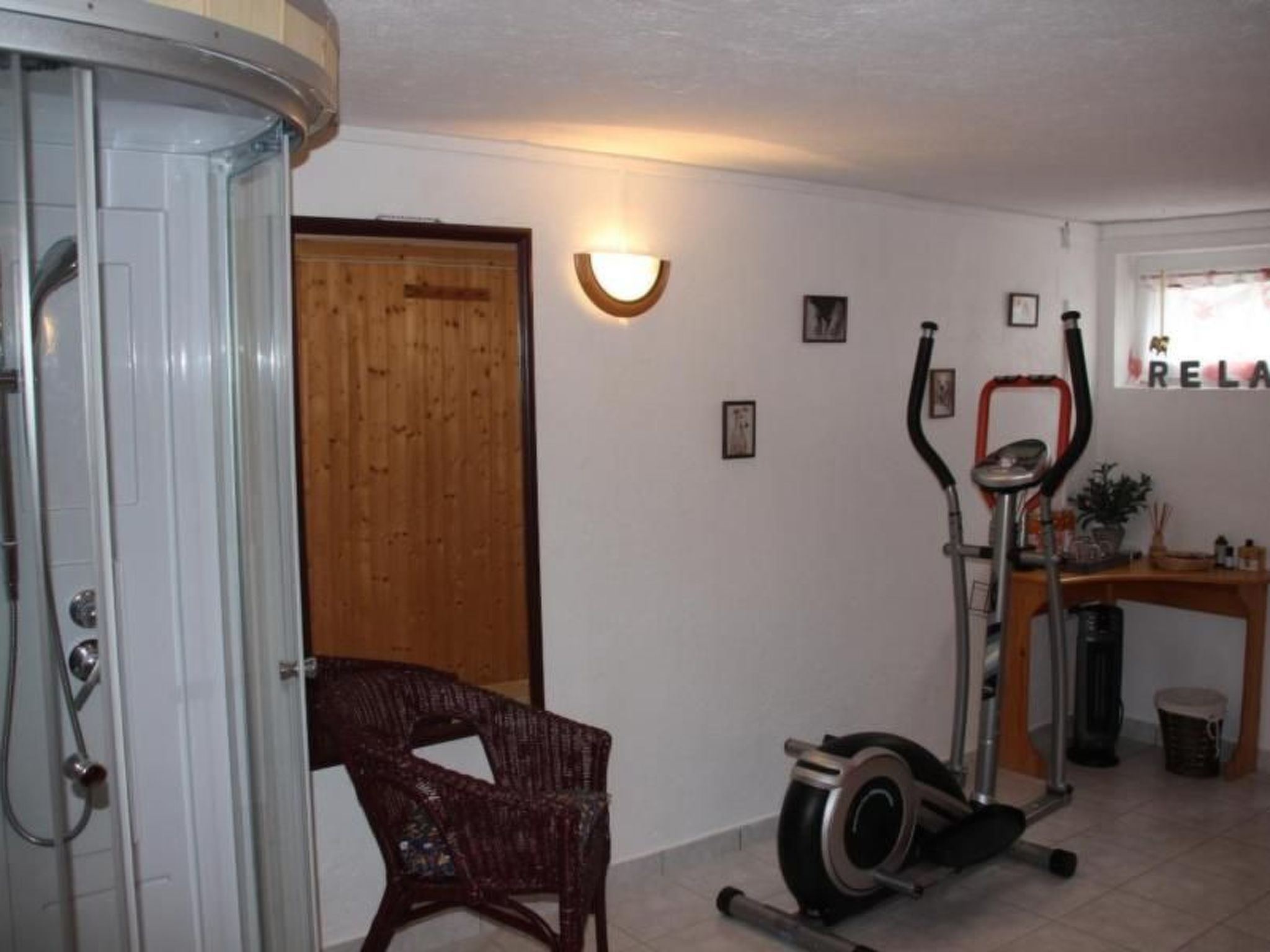 Quarto com porta de madeira, uma cadeira de vime, uma bicicleta ergométrica e uma pequena mesa com uma planta em vaso.