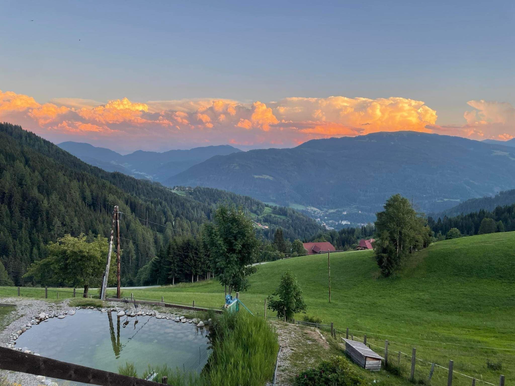 Un paysage rural avec un étang, des champs verdoyants et des montagnes lointaines, le tout baigné par la lumière d'un ciel au coucher du soleil.