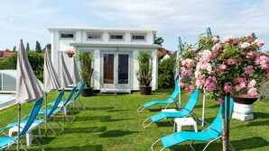Un espace aménagé au bord de la piscine, avec des chaises longues bleues, un bâtiment blanc doté de grandes fenêtres et un pot de fleurs orné de fleurs roses.