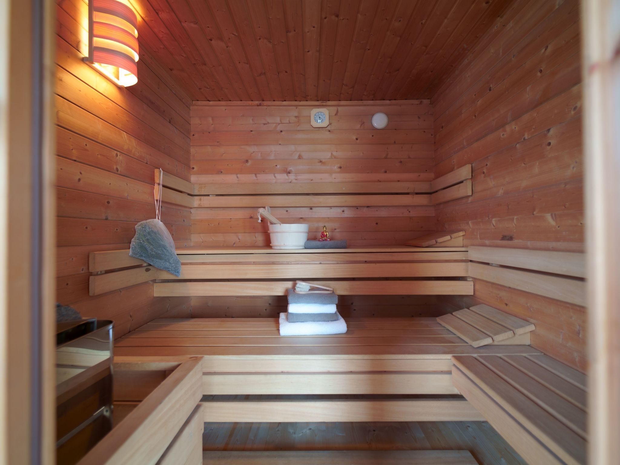 A sauna with wooden benches, a small table with a bucket and brush, and a wall-mounted light fixture.