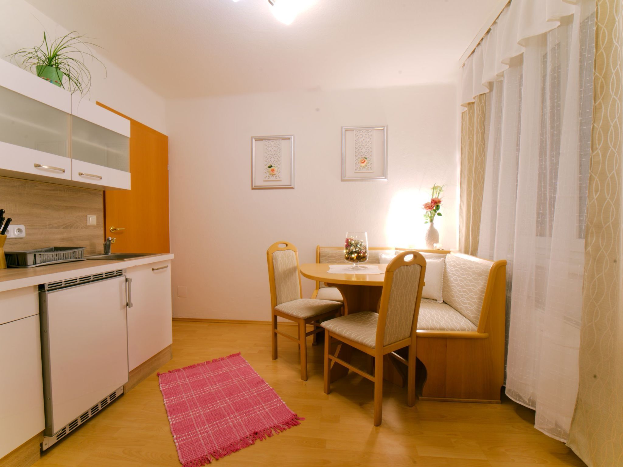 A compact kitchen with white cabinets, a wooden dining table, and two chairs. There is a small rug on the floor and a potted plant on the counter.