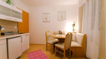 A compact kitchen with white cabinets, a wooden dining table, and two chairs. There is a small rug on the floor and a potted plant on the counter.