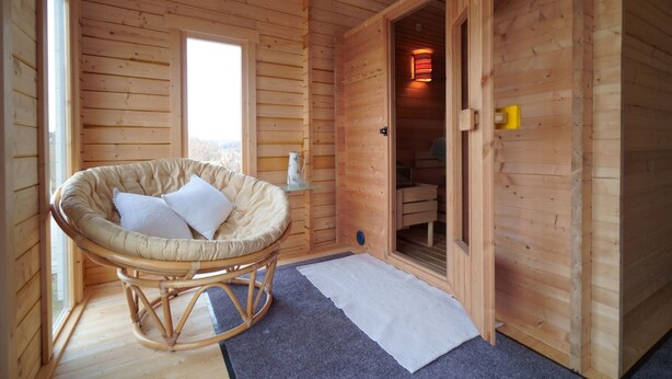 L’intérieur d’un chalet en bois, avec un fauteuil en osier, une petite table sur laquelle se trouve une carafe, et une porte donnant accès à une salle de bain.