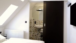 Ein Schlafzimmer mit schrägen Wänden, einem Bett, einem Heizkörper, einem Badezimmer mit Waschbecken und Spiegel sowie einer schwarzen Tür.