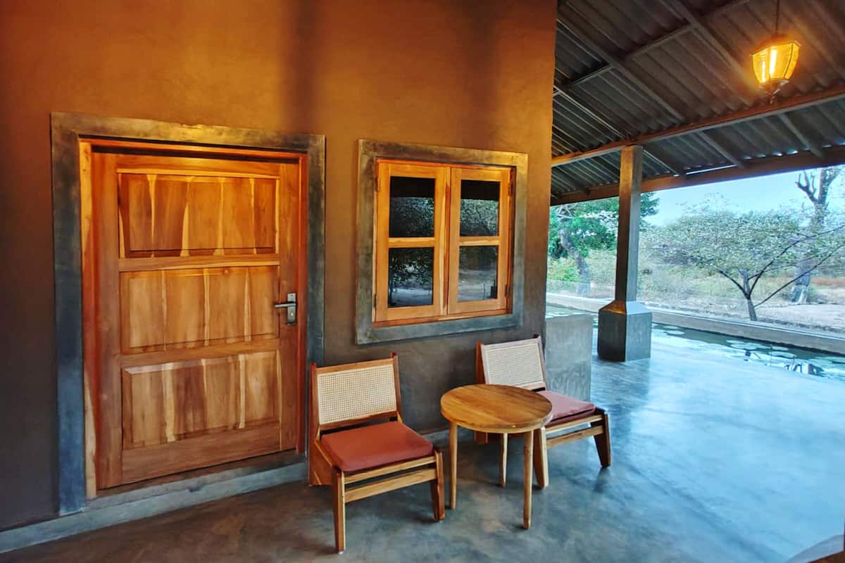 Une porte et une fenêtre en bois, deux chaises et une petite table dans un espace extérieur abrité, avec vue sur les arbres.