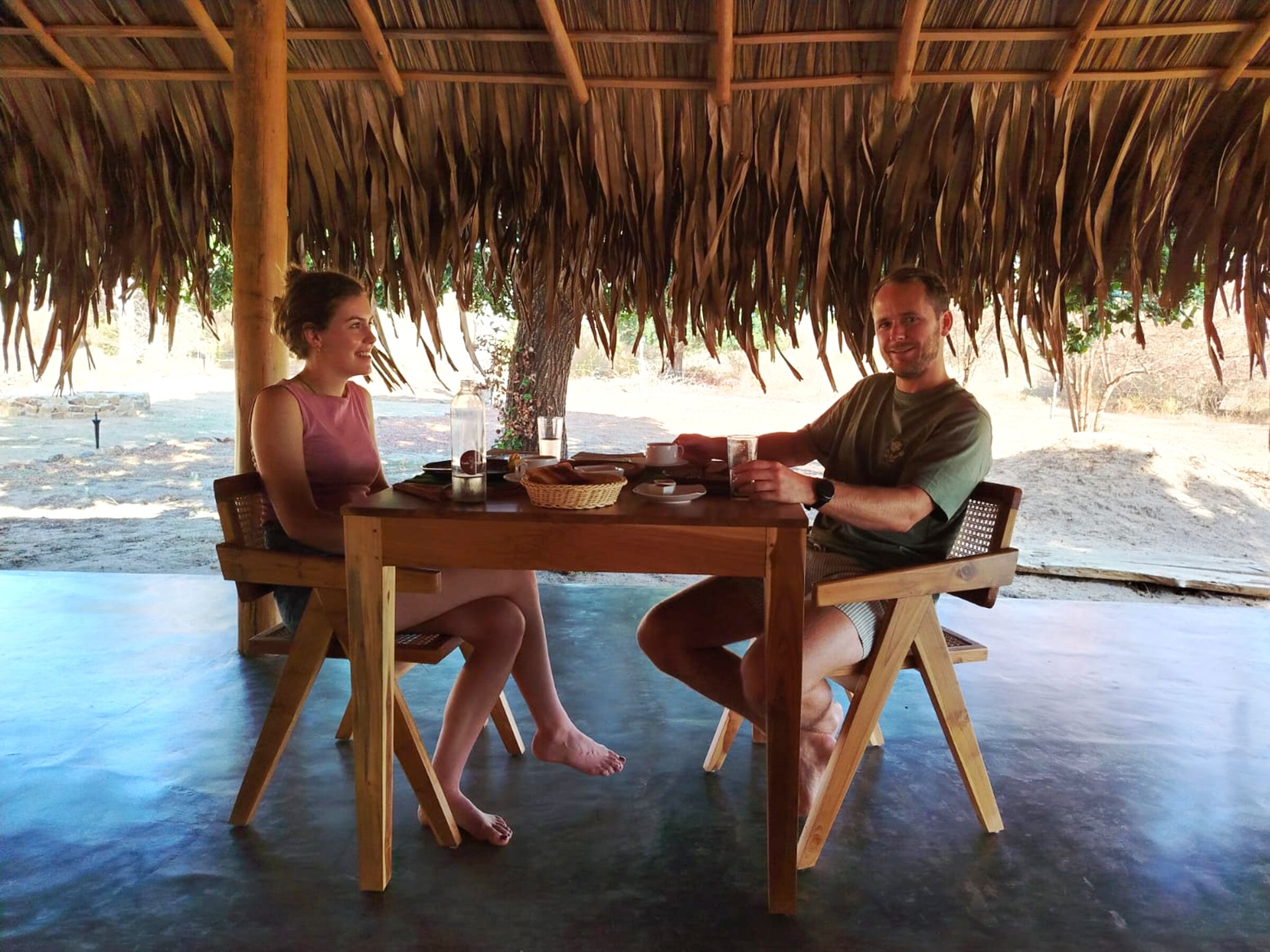 Deux personnes sont assises à une table en bois sous un toit de chaume, avec une vue sur la plage en arrière-plan.