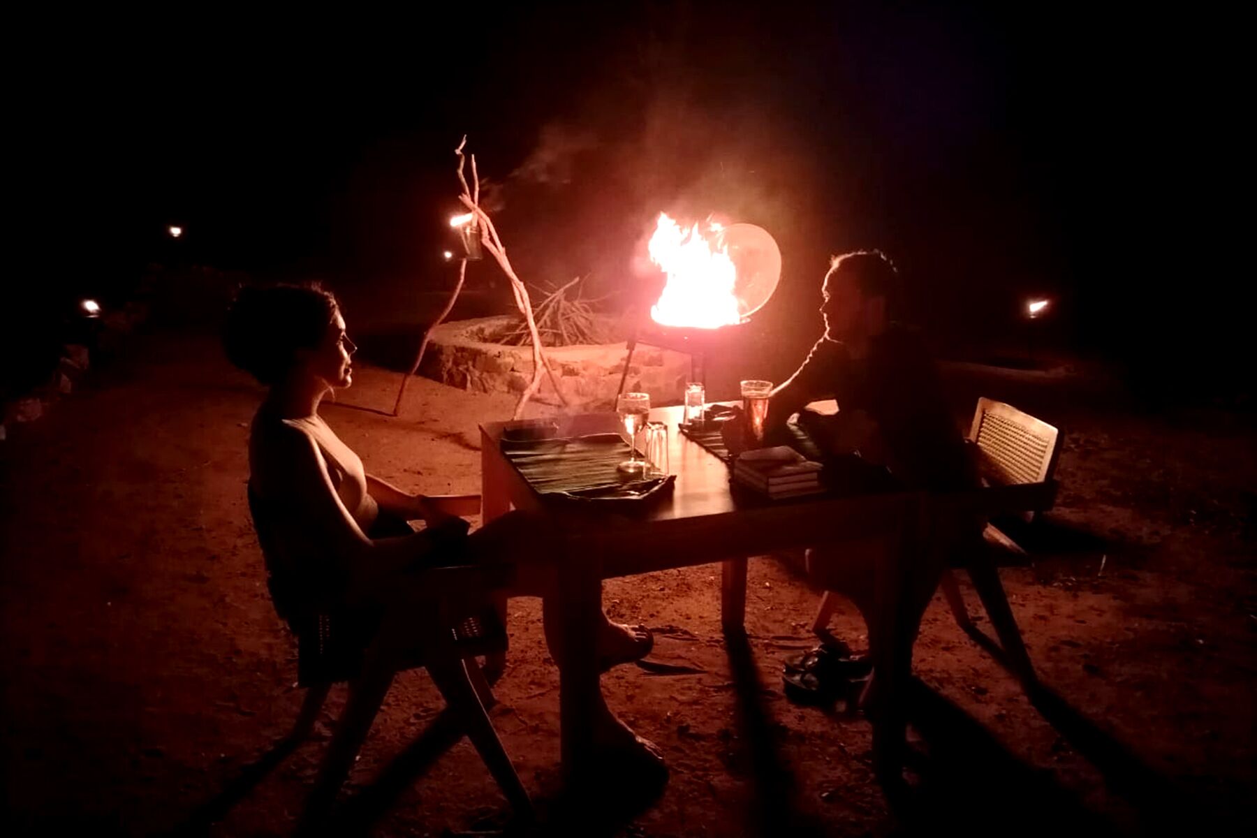 Two people sitting at a table with a campfire in the background.