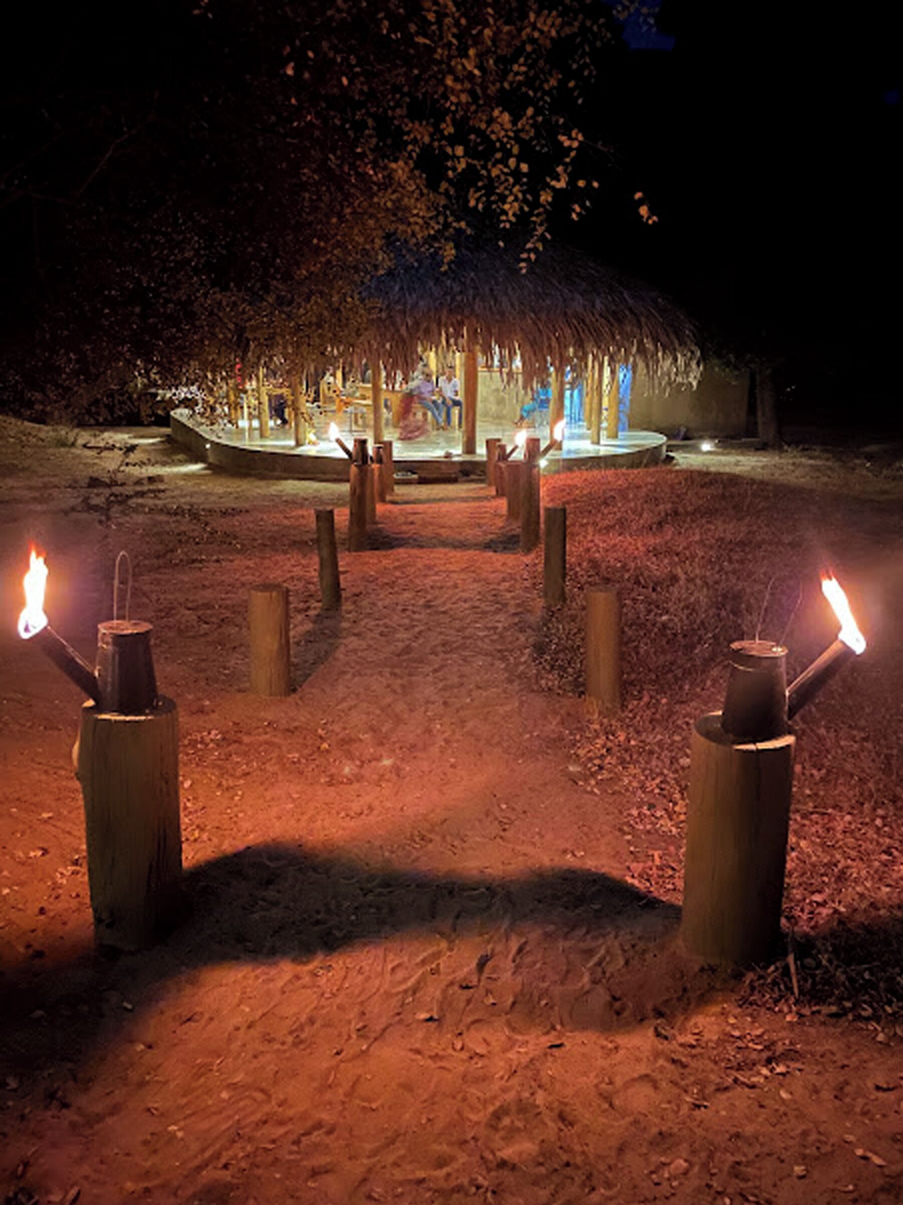 A pathway leading to a thatched-roof structure with a central fire pit, surrounded by wooden posts.