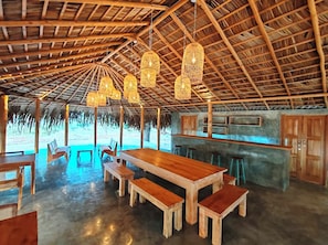 Um restaurante à beira-mar com mesas e bancos de madeira, telhado de palha e luminárias de vime penduradas.