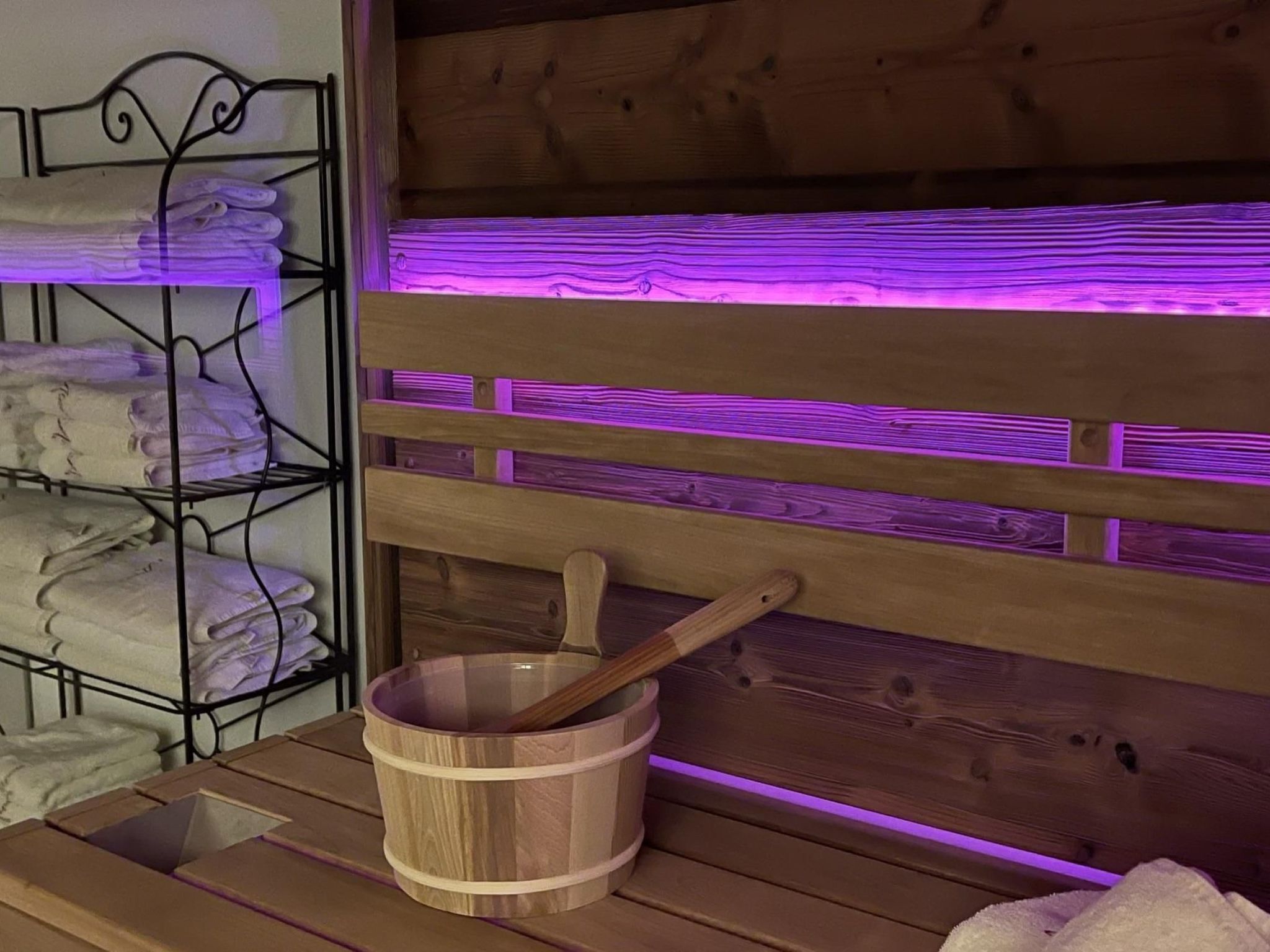 A wooden bench with a wooden bucket and a wooden spoon, a metal shelf with folded towels, and a wooden wall with purple lighting.