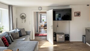 Uma sala de estar moderna com sofá, TV fixada na parede, pequena área de cozinha e corredor que dá para outro quarto.
