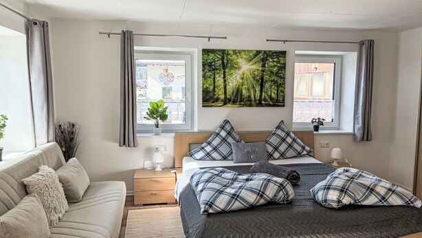 Quarto moderno com cama, sofá, mesa de cabeceira, janela com cortinas e uma obra de arte na parede.